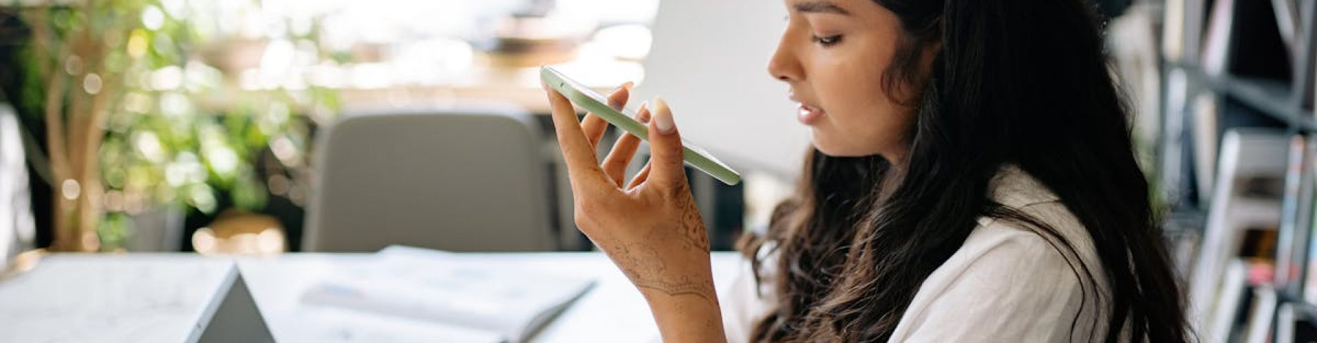 A focused woman uses her phone and laptop in a bright, home office setting.